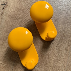 Pode incluir: Dois ganchos de parede amarelo brilhante com a parte superior arredondada e um único gancho na parte inferior. Os ganchos são de plástico e têm um acabamento brilhante.