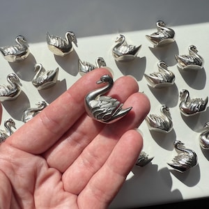 Op de afbeelding: Een verzameling kleine, zilverkleurige zwaanbeeldjes. Eén zwaan wordt in een hand gehouden, met nog veel meer verspreid op de achtergrond. De zwanen hebben gedetailleerde vleugel- en veermotieven en worden op een wit oppervlak getoond.