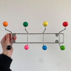 Pode incluir: Um cabideiro multicolorido com oito botões esféricos nas cores laranja, verde, amarelo, vermelho, preto, rosa, azul e verde limão. Os botões são fixados a uma estrutura metálica cromada, projetada para montagem na parede.