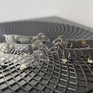 Op de afbeelding: Zes metalen teckel hondjes met zilveren lichamen en goudkleurige poten zijn gerangschikt op een zwart, getextureerd oppervlak. De honden staan in een speelse pose, met hun lange lichamen en onderscheidende kenmerken duidelijk zichtbaar.