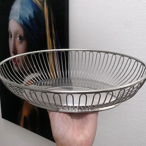 Könnte beinhalten: Eine silberne Obstschale aus Draht mit einer glänzenden, reflektierenden Oberfläche. Die Schale ist oval geformt und hat einen erhöhten Rand.