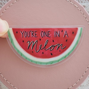 Puede incluir: Una pegatina transparente con un diseño de rodaja de sandía. La sandía es roja con semillas negras y una cáscara verde. La pegatina dice "You're one in a melon".