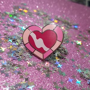 May include: A heart-shaped enamel pin with a pink and red design and a white wavy line. The pin is set against a sparkly pink background with star-shaped confetti.