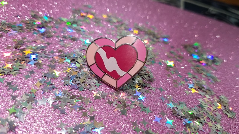 May include: A heart-shaped enamel pin with a pink and red design and a white wavy line. The pin is set against a sparkly pink background with star-shaped confetti.