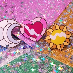 May include: Three enamel pins: a crescent moon in white and lavender, a pink heart, and a yellow sun. Each pin has a gold outline and a white wavy design. The pins are on a glittery background.