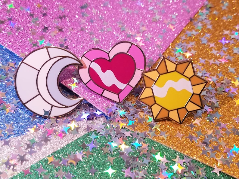 May include: Three enamel pins: a crescent moon in white and lavender, a pink heart, and a yellow sun. Each pin has a gold outline and a white wavy design. The pins are on a glittery background.