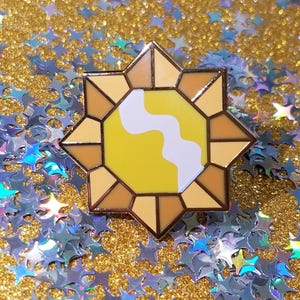 May include: A star-shaped enamel pin with a yellow and white design. The pin has a gold outline and is set against a background of gold glitter and blue star-shaped confetti. This decorative pin is suitable for jackets, bags, or hats.