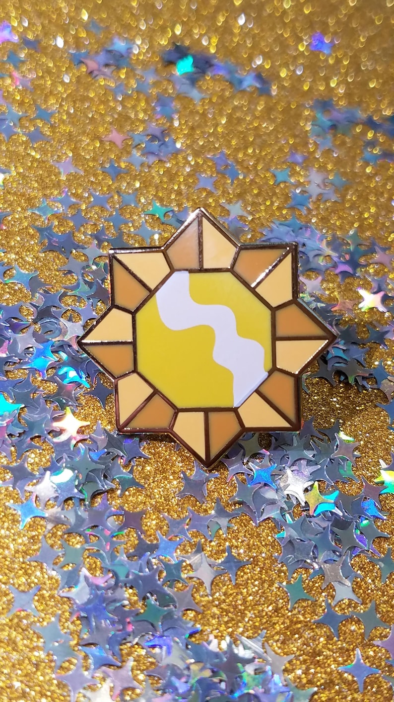 May include: A star-shaped enamel pin with a yellow and white design. The pin has a gold outline and is set against a background of gold glitter and blue star-shaped confetti. This decorative pin is suitable for jackets, bags, or hats.