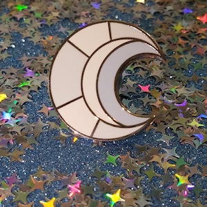 May include: A white crescent moon enamel pin with gold trim. The moon is segmented with black lines. The pin is set against a background of blue glitter and star-shaped confetti.