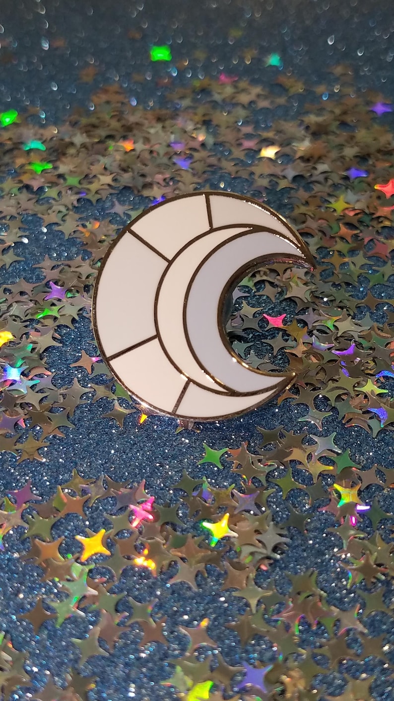 May include: A white crescent moon enamel pin with gold trim. The moon is segmented with black lines. The pin is set against a background of blue glitter and star-shaped confetti.