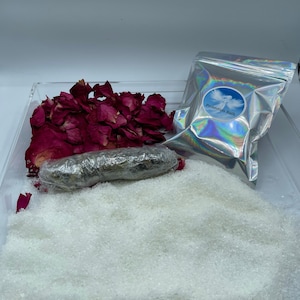 May include: A clear acrylic tray contains bath products. There are dried rose petals, a bundle wrapped in plastic, a bag with the text "Protection Bath", and a large amount of white bath salts. The bag is silver and has a blue and white label.