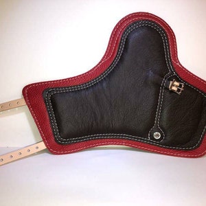 Pu&ograve; includere: Un ginocchiere in pelle rossa con un inserto in pelle nera e dettagli di cucitura bianca. Il ginocchiere ha due cinturini con fibbie.