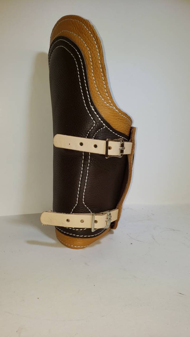 Pu&ograve; includere: Un'imbracatura per lo stinco in pelle marrone con dettagli in pelle color cuoio e cuciture bianche. L'imbracatura ha due cinturini regolabili con fibbie.