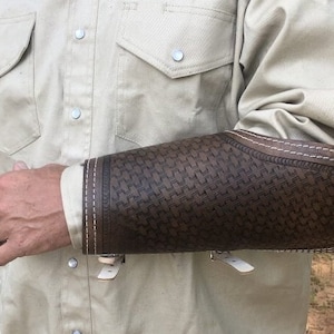 May include: A brown leather arm guard with a white stitched border and a geometric pattern. The guard is worn on the left arm of a person wearing a light beige shirt.