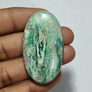 May include: An oval-shaped cabochon gemstone featuring a polished surface with a marbled pattern of deep forest green, light mint green, and white streaks, accented by subtle hints of golden-brown and soft purple mineral inclusions throughout.