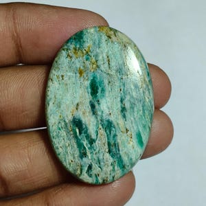 Peut inclure: Une pierre précieuse polie de forme ovale présentant un motif marbré dans des tons de bleu sarcelle vif, de vert clair et de blanc, avec des inclusions brun doré dispersées sur toute la surface lisse et réfléchissante de la pierre.