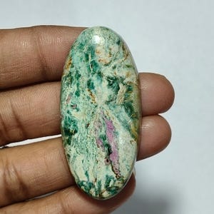 Peut inclure: Une pierre précieuse polie de forme ovale présentant un motif marbré de vert forêt profond, de blanc crème et de brun clair, avec une tache allongée distincte de rose magenta vif courant verticalement au centre de la surface.