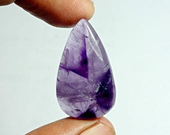 May include: A teardrop-shaped amethyst gemstone, showcasing a blend of deep purple and lighter lavender hues. The polished surface reveals natural patterns and inclusions, held between two fingers against a white background. The gemstone is a beautiful example of natural crystal formations.