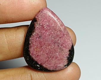 Könnte beinhalten: Ein tropfenförmiger Edelstein mit rosa Mitte und schwarzem Rand. Der rosa Bereich hat ein strukturiertes Aussehen. Der Edelstein wird zwischen zwei Fingern gehalten.