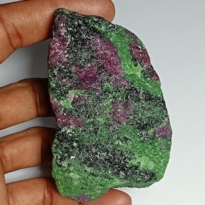 Peut inclure: Une pierre précieuse brute de rubis dans de la zoïsite. La pierre présente un mélange de rubis rouge foncé, de zoïsite verte et d'accents noirs. La pierre naturelle est tenue dans une main sur fond blanc.