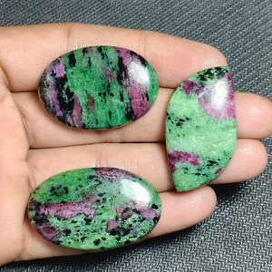 May include: Three polished ruby zoisite gemstones. The stones have a mix of green, pink, and black colors. Two are oval-shaped, and one is a half-moon shape. The gemstones are held in a hand.