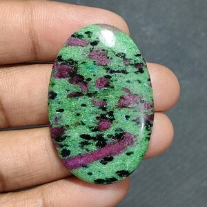 Può includere: Una pietra preziosa lucidata di forma ovale con un colore di base verde vibrante, inclusioni magenta-rosa intenso sparse e piccole macchie nere su tutta la superficie, caratterizzata da una finitura a cabochon liscia e riflettente.