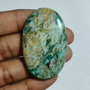 Peut inclure: Une pierre précieuse polie de forme ovale présentant un motif marbré de bleu sarcelle, de vert forêt et de bleu clair avec des veines brun doré proéminentes et une petite inclusion rose magenta distincte près du bord inférieur, finition lisse.