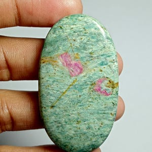 May include: An oval gemstone with a polished surface. The stone has a mottled pattern of light green and blue-green hues, with pink and brown inclusions. The gemstone is held by a hand.