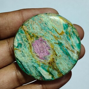 Peut inclure: Un cabochon de pierre précieuse poli et circulaire présentant une surface marbrée vert et bleu sarcelle vibrante avec des stries dorées et une inclusion magenta-rose texturée et proéminente au centre, avec une finition lisse.