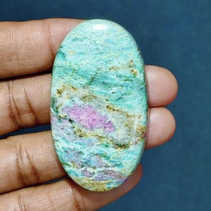 Könnte beinhalten: Ein ovaler Cabochon-Edelstein mit einer polierten Oberfläche, die ein marmoriertes Muster aus hellem Türkis, Minzgrün und Weiß aufweist, akzentuiert durch deutliche Flecken in tiefem Magenta und feine Adern aus dunkelbraunen Mineraleinschlüssen.