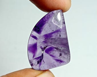 May include: A polished, semi-translucent gemstone with a deep purple hue and a unique, starburst-like pattern. The stone is held between two fingers, showcasing its smooth, curved shape and natural variations. The gemstone is approximately 1.5 inches in length.