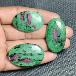 Op de afbeelding: Drie gepolijste, ovaalvormige cabochons met een levendige groene basis en duidelijke insluitingen van diep magentarood en zwarte minerale strepen. Deze gladde, afgeronde edelstenen hebben een glanzende en reflecterende textuur.