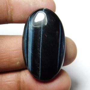 Puede incluir: Cabujón ovalado de ojo de tigre azul con un patrón azul oscuro y blanco. La piedra es lisa y pulida.