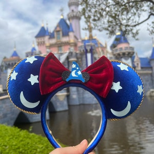 May include: A blue velvet headband featuring two circular ears decorated with white crescent moons and stars. A large, deep red velvet bow sits between the ears, centered with a small blue wizard hat emblem featuring a white moon and star.