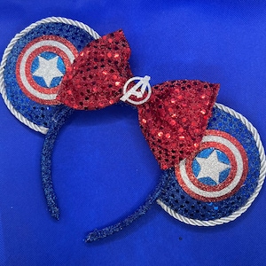 May include: Blue sequined headband with Captain America shield-themed ears and a red sequined bow. The ears have a silver rope trim and a silver star in the center. The bow features a silver Avengers logo.