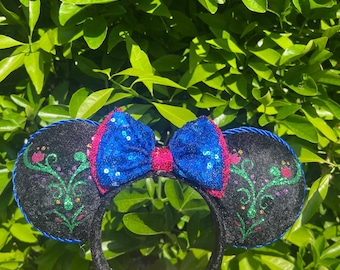 May include: A black headband featuring two round ears with green, pink, and gold floral embroidery. The center includes a bright blue sequined bow with a pink sequined knot. The edges of the ears are trimmed with a blue twisted rope cord.