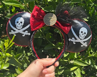 Puede incluir: Orejas de ratón negras con temática pirata. Las orejas presentan un diseño de calavera y huesos cruzados con detalles de purpurina plateada. Un lazo rojo y una moneda de oro con una calavera están centrados en la diadema, con plumas negras.