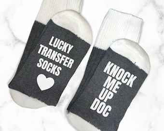 May include: A pair of grey and white socks with the text "Lucky Transfer Socks" and a heart on one sock and "Knock Me Up Doc" on the other sock.