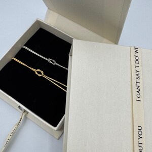 May include: Two delicate knot bracelets, one gold and one silver, are displayed in a white box with a black interior. The box is open and the bracelets are visible. The box has a white ribbon with the text "OUT YOU I CAN'T SAY 'I DO' WITH" printed on it.