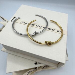May include: Two gold and silver knot bracelets in a white box with the text "I can't say 'I do' without you".