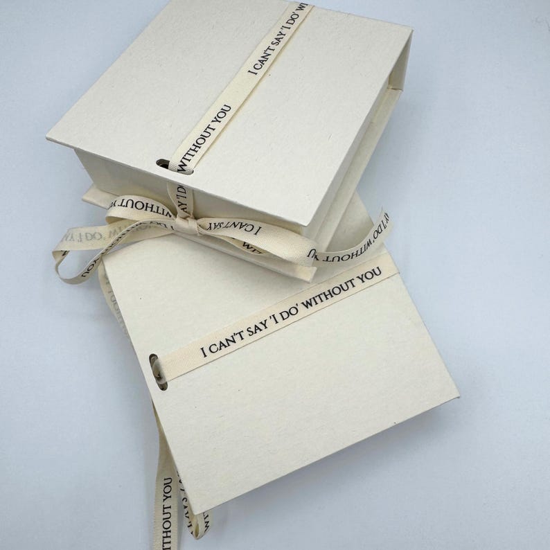 May include: Two cream-coloured boxes tied together with a cream ribbon. The ribbon has the text "I can't say 'I do' without you" printed on it.