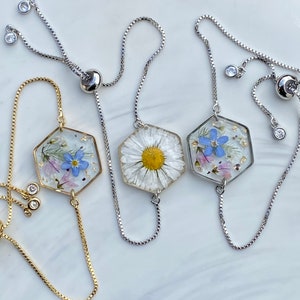 May include: Three adjustable bracelets with hexagonal charms featuring pressed flowers. The charms are made of clear resin and contain a daisy, forget-me-nots, and a mix of wildflowers. The bracelets are made of silver and gold chains.