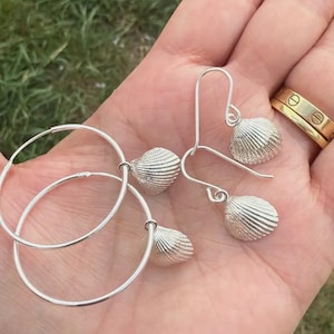 May include: A set of silver-toned jewellery consisting of two hoop earrings and two drop earrings. Each earring features a small, textured seashell charm with distinct ridges. The hoop earrings are circular, while the drop earrings hang from curved hooks.