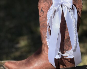 May include: A brown leather cowboy boot with decorative stitching. A white satin bow and a white lace garter with dangling ribbons are attached to the boot, suggesting a wedding theme.
