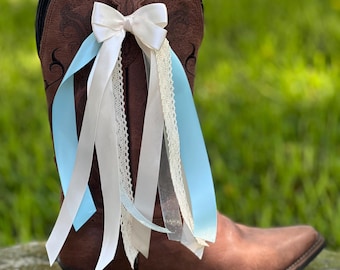 May include: A brown leather cowboy boot adorned with a white bow and flowing ribbons in light blue, white, and cream. The boot has decorative stitching and a pointed toe. The ribbons add a touch of elegance to the rustic footwear.