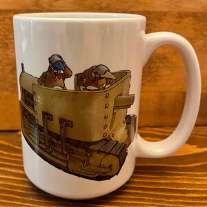 May include: White ceramic mug with a cartoon illustration of two figures in a tank. The tank is brown and has a black track. The figures are wearing blue hats and red bow ties.