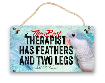 May include: A teal blue wooden sign with a white bird and the text "The Best Therapist Has Feathers And Two Legs". A blue and gray parakeet is on the right side of the sign.