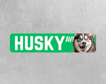 May include: A green street sign with the text "HUSKY AVE" in white. A smiling husky dog is on the right side of the sign. The dog has black and white fur and blue eyes. The background is a light gray.