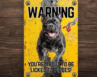 May include: A rectangular metal sign with a yellow background and black text that reads "WARNING" above a black dog. The text "YOU'RE ABOUT TO BE LICKED TO PIECES!" is below the dog. The sign has a distressed look.