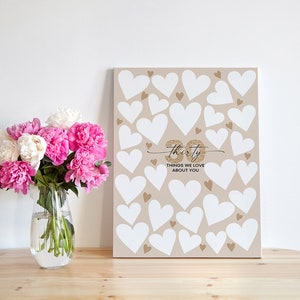 May include: A beige canvas sign with white hearts and gold glitter. The sign says "Thirty Things We Love About You".